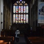 The nave of a church with a woman standing in it, and a large stained-glass window.