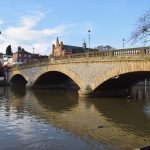 A yellow-stone bridge across a river, with a tree to the left and some buildings in the background.