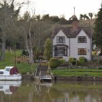 A house and some trees across a river, with a motorboat and a chain ferry in the water.
