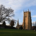 A tree and church-style tower, with a church and spire in the background, and grass in the foreground.