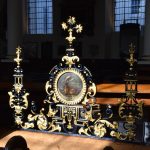 Ironwork screen, partly gilt and partly painted black, with two side pillars and a central section containing a painting of a stag on a field of grass. Behind are windows and pews.