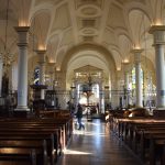 The interior of a cathedral. White pillars on either side of a central aisle lead to and behind an ironwork screen. The simply vaulted ceiling is white with gold decoration. Wooden pews lead up to the screen. There are two stained-glass windows in the background, with windows of plain glass on the side walls.