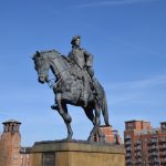 A dark metal equestrian statue of Bonnie Prince Charlie, on a stone plinth, against a blue sky with contrails. Towards the bottom of the frame are buildings of various ages, all of red brick, with bare trees.