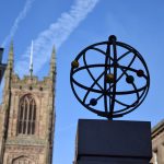 An atom-like metal structure, with coloured balls around the outside and a golden nucleus, on a square stone plinth. Behind it are a church tower and various other buildings, all in soft focus.
