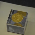 A gold coin with an intricately embossed image of a ship, on a small glass display cube.