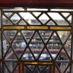 A leaded window, with “X” patterns, as well as some horizontal and vertical thin rectangles stained orange, in a brown frame. Behind it, in soft focus, are a collection of shelves containing vases and other objects, around the edge of a balcony overlooking another space. The room behind the window is lit from above.