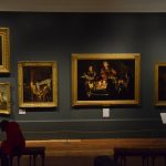 A green wall, with a white crown moulding, against a light wooden floor. On the wall hang seven paintings in gold frames, two cut off on the right, many depicting candlelit scenes. There are white information plates beneath the pictures, and a further one on its own stand. In the foreground are two wooden benches; a person it sitting and reading on the left-hand bench.