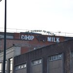 Looking up to the tops of buildings, of stone and brick, of various ages. The building furthest back carries a sign. The sign reads, firstly, “THE GATEWAY TO HEALTH”, in upper-case dark green brush script font, in an arch shape. Beneath that are the words “CO-OP MILK”, in white upper-case sans-serif, between which is an image of a brown cow (with ears, eye and udders detailed).