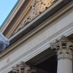 In light stone, the tops of Corinthian columns, above which is an entablature bearing two roses on squares, between which is engraved “MCMXLI”. Above all is a pediment, containing a coat of arms. In front flies a flag with a white background, also bearing a coat of arms.