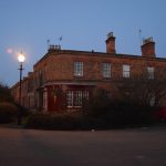 Lit by an gas-style streetlamp in twilight are red-brick cottages on a street corner. At the corner is a former shop. On the edge of the pavement is shrubbery, to the right of which is a parked car, the road being completely clear.
