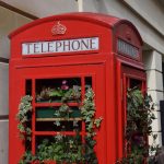 A red telephone booth against a light stone wall, with recesses for windows and minor detailing. The telephone booth lacks glass, and there are plants inside protruding through the frame. Some people are in the background.