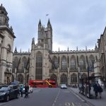 Looking down a street. There is a stone civic building to the left and stone shops to the right. At the far end is a large church, with high gothic windows. The transept contains a clock, while from the central tower flies a Union Flag. A red bus is parked in front of the church.