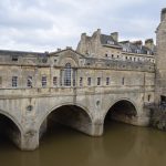 A stone bridge crosses a river. The bridge has three arches, and atop those a high wall with windows (one large at the centre) and a roof. The far side of the bridge leads to more buildings. The sky is overcast, and the river water a murky brown.