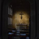 A dimly-lit chapel in a church, seen through its doorway. A circular area of the wall is illuminated, where an elaborate cross hangs on a wall with an altar below it. Simple chairs face the altar.