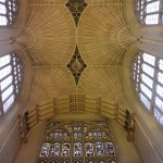 Looking up at the ceiling of an abbey. Three walls are visible, the centre one containing a large stained-glass window, the other two containing two of plain glass. Fan faulting rises from the columns. There is some painted detailing on the ceiling.
