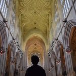 Looking down the nave of an abbey, with gothic arches to either side and windows above. There is fan vaulting on the ceiling. The view is low, and the east end of the abbey is partially obscured by a man’s silhouette.
