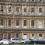 A wall of Georgian townhouses, with sash windows between different classical orders of columns. The houses have three stories with a roof above, and are of light stone, with white doors and window frames. There are parked cars partially obscuring the lowest tier, and five people in two clusters around two of the doors.