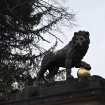 A stone lion on a gate post, three of its paws resting on the plinth with the fourth on a golden ball. The plinth also supports two smaller golden leaves, one partially out-of-frame. There is a tree in the background.