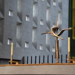 On a table stand two candles in simple candlesticks, and a formed metal cross containing two crossed nails at its centre. A wall with small and large windows, and a large tapestry, form the background.