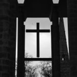 Black-and-white photo. A cross is suspended from a grid of concert pillars. In the background are bare trees and a church spire.