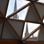 Two church spires are visible through two offset square panes of glass, with opaque squares in between to form a chequered pattern. Criss-crossing the whole diagonally is a wooden frame.
