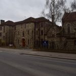 A flint-stone building, three storeys high, with a dark roof, and a chapel of the same materials to the side. The building stands on a quiet road, with a lamppost, bus stop and bikes near the background.