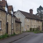 Light-stone buildings of two storeys with red roofs. One has dormer windows. The buildings are on a road with a partial grass verge, which winds into the distance.