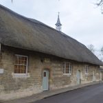 A building with front wall of light stone and a side wall of brick, with four sash windows and three pale green doors in the front wall. The building has a thatched roof as high as the wall, topped with a small bell tower and spire.
