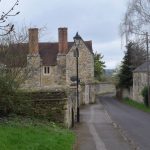 A street with lamppost. There is a grass verge on the left in the foreground, and a large light stone house with red roof and high brick chimney stacks behind. In the distance the road, flanked by a high wall, curves around to the right.