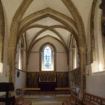 A brightly-lit square space at the end of a church, with wood panelling at the base of the far wall (this also contains a stained glass window). The roof vaults are gothic, with stone ribs and white infill.