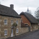 A village street scene, with a stone house with a dark roof in the foreground and a thatched church hall (of stone and brick) behind, both on the left of the street. There are bare trees in the background.