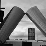 Black-and-white photo. Two metal arches lean against each other, framing a low building and a high-rise tower against a cloudy sky.