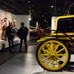 A yellow and black vintage car is half-visible in the foreground. In the background are other displays or cars and costumes, while two visitors walk past.