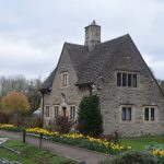 A two-storey house of grey stone, in a turfed garden. A path runs across the front, with yellow flowers between the path and the garden; a person stands on the path. In front of the path is a small grass slope, with stone steps down to the side of a canal (the water itself is out of shot).