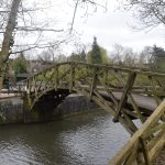 A geometric wooden footbridge over a canal, leading to some squat buildings. All around are trees, with buildings just visible in the background.