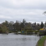 A canal, seen from the towpath on the right. There is an island in the canal, with a stone bridge leading across to it from the towpath. A house sits on the island, and there are more buildings in the background on the left bank. Chains hang across the canal blocking access to the path to the left side of the island.
