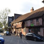 Half-timbered buildings, with the part in between the timbers pink, and thatched roofs. Behind are concrete buildings. There are modern signs on the buildings, and modern cars (and people) in the street.