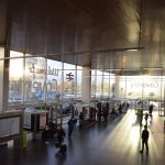 A large atrium with glazed walls, and light pouring in. Information boards stand in the hall, along with people waiting around. Outside the windows are concrete buildings and cars.