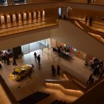 Looking down into an atrium, with staircases crossing it. The floors are of food on the balconies and tile on the bottom. There are people milling around the bottom and a few on the balconies. There are also entrance doors at the bottom, along with a racing car, an entrance desk and a shop.