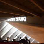 A curved concrete roof, with straight beams supporting it which curve around the picture. Light pours in from a series of triangular windows, illuminating part of the roof. In front of the illuminated part is a silhouette of an old car.