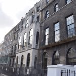 A dark brick building with a central portion made of lighter stone, s part of a terrace of four- and five-storey buildings. There is a lamppost, and an overcast sky.