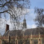 A large red-and-yellow-brick building, with high narrow gothic windows, castellated walls, and a spire and chimneys atop the high roof. In front are bare trees, against a lightly-clouded sky.