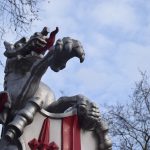 A close-up of a dragon sculpture, standing on its hind legs (not visible). Visible are its head, upper body and front legs, and part of a wing. One front leg is raised, the other holds a shield, the top part of which is visible (a white background, with a red sword and a red vertical stripe). The dragon itself is painted silver, with a red tongue and red detailing on its wing. In the background is a blue sky with clouds, and portions of bare trees.