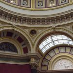 An elaborately decorated upper wall, and part of ceiling. Arches are seen in a line along a curved wall (two in full, two partially), with one of the central ones containing a bronze relief and one a recessed section with a skylight, leading to an arched wall with more decoration and a marble relief. The walls are painted in red, with cream near the top, and red, blue, green and gold detailing. The lower portion of a dome is seen, with white flowers in blue squares forming the detailing there.