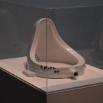 The artwork “Fountain”—a urinal, displayed on its side and painted white, signed “R. MUTT 1917” in black pen—sits inside a glass case on a grey plinth.
