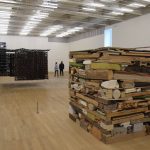 A quiet, large, art gallery, with light wooden floors and white walls. Two large installations are in the centre: a perfect cube made from various materials (wood, card, fabric etc) in a pile, and a dark hanging in the form of walls. Other artworks are visible on the far walls.
