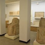 A museum gallery. In the centre of the picture two square pillars, to the right of each of which is a seated stone figure, heavily weathered and with the head missing in each case. Behind, on shelves (some supported by brick) are stone tablets on the walls. The floor is of dirty-looking tile, and there are office-style ceiling tiles.