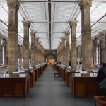 A large room, with two sets of columns symmetrically placed leading into the distance, between which are identical wooden museum cabinets. The walls are of grey and yellow stone, and the ceiling is white with a plaster decoration.