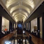 A long, narrow gallery, seen from the centre. There is a table and a cabinet on the plinth in the centre. The roof is vaulted, with skylights; the floor wooden. The walls are black, with white recesses containing various items of furniture grouped by use (e.g. chairs).
