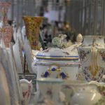 A shelf of porcelain, seen from the side, so the items (vases, teapots, cups) stretch into the distance. The porcelain is highly decorated, mostly white, some gilded. Much of it is out of focus.